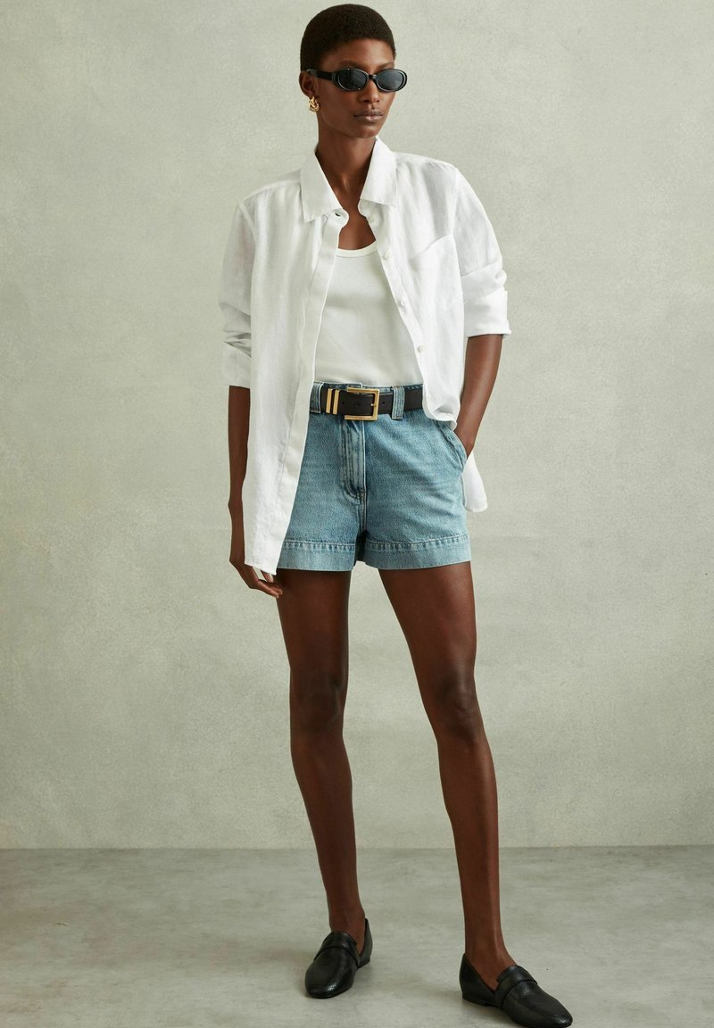 Hvid linned skjorte, tætsiddende hvid tanktop, lyseblå denim shorts, sort bælte med guldfarvede accenter, sorte loafers og sorte solbriller.