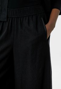 Pantaloni neri in lino con cintura elastica e tasca laterale, caratterizzati da una finitura testurizzata sottile e elementi di design minimalisti.