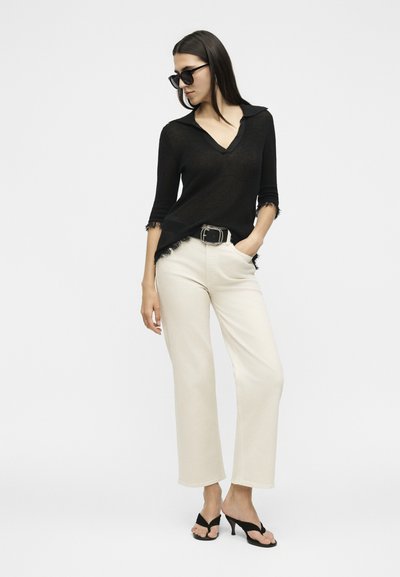 Γυναίκα με μαύρο τοπ V-neck με φθαρμένες άκρες, εκρού φαρδιά παντελόνια με μαύρη ζώνη, μαύρα σανδάλια με τακούνι και σκούρα γυαλιά ηλίου, όρθια.