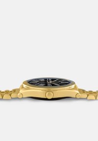 Gold wristwatch with a round black dial, textured hour markers, and a link bracelet. Features a prominent crown and black accents.