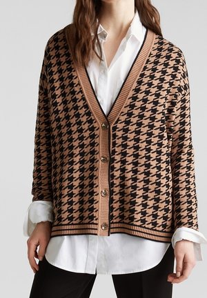 Femme portant un cardigan boutonné marron et noir à motif pied-de-poule sur une chemise blanche avec un pantalon noir, vue des épaules aux hanches.