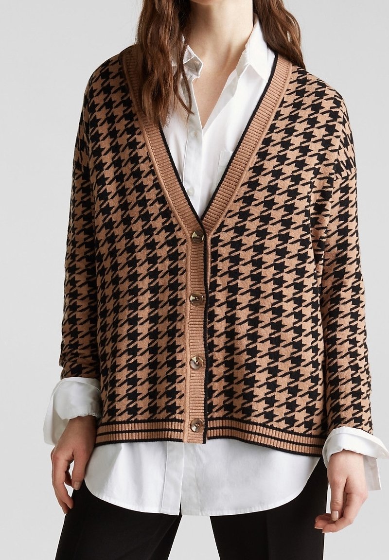 Femme portant un cardigan boutonné marron et noir à motif pied-de-poule sur une chemise blanche avec un pantalon noir, vue des épaules aux hanches.