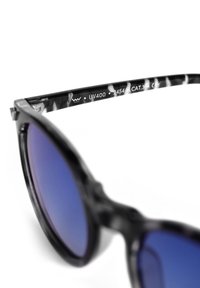 Sluneční brýle s černobílým vzorem a modrými skleněmi na vnitřní nožce s informacemi o UV400 a modelu.