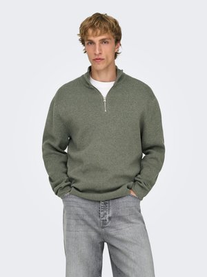 Jovem com cabelo claro e ondulado veste um suéter verde com fecho até meio por cima de uma camisa branca e jeans cinza claro, com as mãos nos bolsos, em pé contra um fundo simples.
