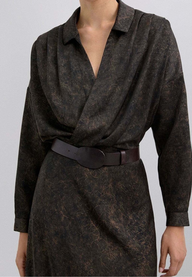 Femme portant une robe portefeuille sombre et texturée avec des manches longues et une large ceinture noire avec une boucle ovale à la taille.