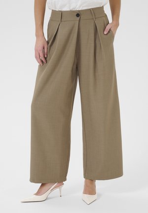 Pantaloni beige a gamba larga realizzati in tessuto testurizzato, con pieghe frontali, chiusura con bottone e tasche laterali. Indossati con scarpe con tacco alte bianche.