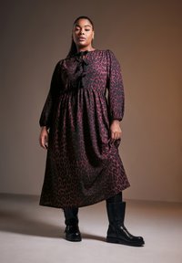 Mekko, jossa on musta ja burgundinruskea leopardikuosi, pitkät hihat, pyöreä pääntie ja nappiyksityiskohtia, yhdistetty mustiin polvipitkiin saappaisiin.