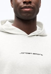 Κλείσιμο σε πλάνο ενός ατόμου που φοράει ανοιχτό γκρι φούτερ με κουκούλα, με το κείμενο "Jordan Sport" τυπωμένο με μαύρα γράμματα στο στήθος.