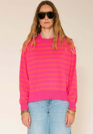 Femme portant de grandes lunettes de soleil noires, des boucles d'oreilles en or, un pull rayé rose et rouge, un jean bleu clair et des bracelets en or.