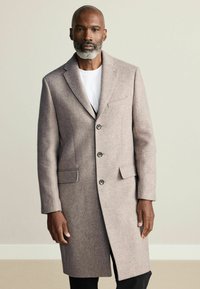 Długi, dopasowany płaszcz w jasnobrązowej wełnianej mieszance. Ma kołnierz z wycięciem, trzy guziki i dwie przednie kieszenie. Gładka faktura, nowoczesny design.