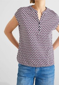 Blusa a maniche corte con scollo a V, caratterizzata da un motivo geometrico in viola e bianco, dettagli con bottoni e vestibilità morbida. Realizzata in tessuto leggero.