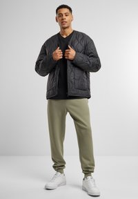 Veste bomber noire matelassée avec fermeture éclair à l'avant, associée à un pantalon de jogging olive et des baskets blanches. Le modèle se tient devant un fond neutre.