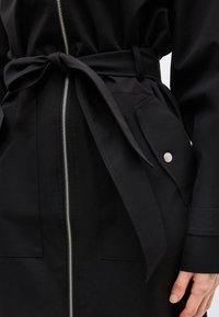 Czarny strój z przednim zamkiem błyskawicznym, wiązanym paskiem z tkaniny, dużą klapą kieszeni z zatrzaskiem i widoczną dłonią z boku.