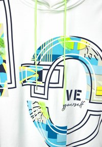 Sudadera blanca con patrones geométricos en azul, amarillo y verde. Presenta contornos negros y un cordón en verde neón. El texto dice "ÁMATE a ti mismo."