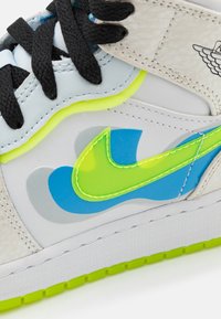 Bílé tenisky s texturovanou panelem, světle modrými a šedými akcenty, jasně zeleným Nike swoosh, černými šňůrkami a neonově žlutým lemem na mezipodešvi.