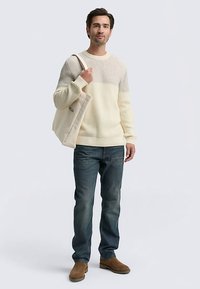 Hellgrauer und cremefarbener gerippter Pullover mit Streifen, blaue Jeans, braune Wildlederstiefel, hält eine beige Tragetasche mit Logo. Stehende Pose.