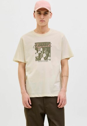 Beige Baumwoll-T-Shirt mit kurzen Ärmeln, das eine Kaktusgrafik in Dunkelgrün und rosa Akzenten zeigt. Kombiniert mit dunkelbraunen Hosen.
