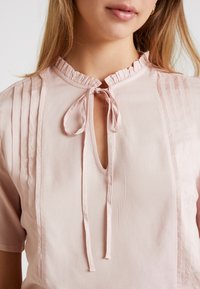 Joven mujer vestida con una blusa rosa claro con cuello con volantes, abertura en el pecho y detalle de moño atado, mostrando la parte superior del torso y los labios.