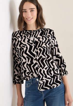 Langärmlige Bluse mit schwarz-weißer abstrakter Musterung, ausgestattet mit einem runden Kragen und einer Knopfleiste, kombiniert mit blauen Jeans.