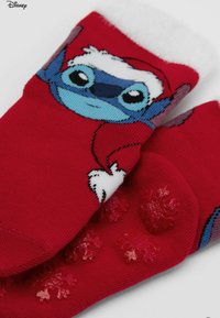 Calze di cotone rosso con un personaggio dei cartoni animati blu che indossa un cappello da Babbo Natale. Polsino morbido bianco e dettagli antiscivolo in silicone a forma di fiocco di neve sulla suola.