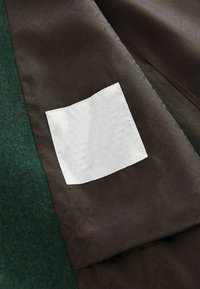 Dunkelbrauer Stoff mit einem subtilen Blumenmuster; daneben ist die grüne Wolltextur sichtbar. Eine weiße quadratische Tasche ist auf den dunklen Stoff genäht.
