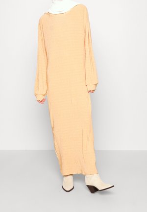 Długa, luźna sukienka w kolorze beżowym wykonana z fakturowanej tkaniny, z długimi rękawami, szerokim dekoltem i marszczonymi mankietami. W zestawie z białymi botkami za kostkę.