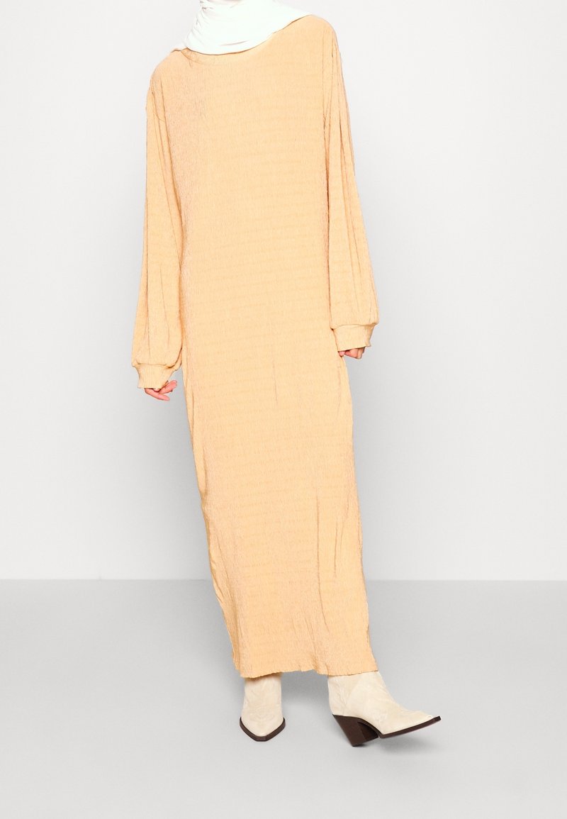 Robe beige longue et ample en tissu texturé, à manches longues, avec un large décolleté et des poignets froncés. Associée à des bottines blanches à la cheville.