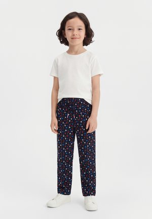Enfant aux cheveux foncés mi-longs, portant une chemise blanche à manches courtes, un pantalon fleuri bleu marine et des baskets blanches, debout devant un fond blanc.
