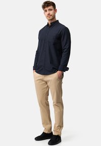 Camisa de botones azul marino con un bolsillo en el pecho, combinada con pantalones beige de corte slim y zapatillas negras. Tejido suave y corte ajustado.