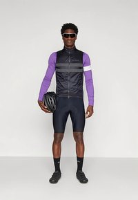 Άνδρας ποδηλάτης με μοβ μακρυμάνικη φανέλα, μαύρο χωρίς μανίκια γιλέκο, μαύρες ποδηλατικές βερμούδες, γυαλιά ηλίου, κρατώντας μαύρο κράνος, στέκεται μπροστά σε λευκό φόντο.