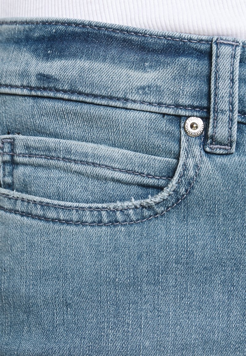Gros plan sur la poche arrière d'un jean en denim bleu clair avec passants de ceinture et rivet métallique sur fond en tissu blanc.