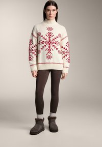 Cremefarbener Pullover mit rotem Schneeflockenmuster, hohem Kragen und gerippten Bündchen, kombiniert mit braunen Leggings und grauen Knöchelstiefeln mit Fellbesatz.