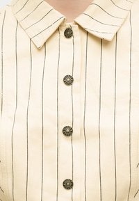 Μπεζ πουκάμισο με κουμπιά, με λεπτές μαύρες κατακόρυφες ρίγες, μυτερό γιακά και μεταλλικά κουμπιά χαραγμένα με το κείμενο "GUESS JEANS".