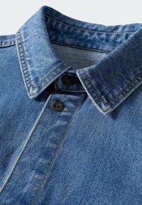 Col de chemise en denim bleu avec des coutures visibles et deux boutons noirs sur la patte de boutonnage, montrant la texture et les détails du tissu.