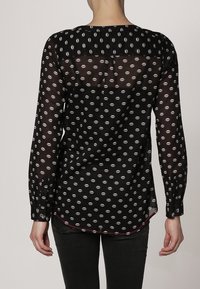 Blouse noire à manches longues avec imprimé de lèvres blanches, tissu transparent, encolure ronde, ourlet légèrement arrondi, et poignets à boutons.