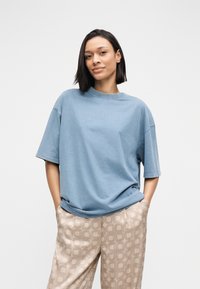 Φαρδιά βαμβακερή μπλούζα σε ανοιχτό γαλάζιο χρώμα με κοντά μανίκια και στρογγυλή λαιμόκοψη, συνδυασμένη με μπεζ παντελόνια με γεωμετρικά σχέδια.