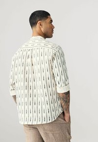 Chemise à manches courtes couleur crème avec des motifs verticaux noirs. Dotée d'un col boutonné et de manches roulées pour un ajustement décontracté.