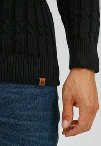 Pull en maille noire avec des poignets et un ourlet côtes, featuring une petite étiquette en cuir brun en bas. Porté sur un jean en denim.
