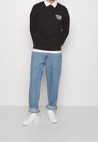 Maglione nero con logo "TOMMY JEANS" sul petto, camicia bianca con colletto sotto, jeans azzurri chiari e sneakers bianche con punta arrotondata.