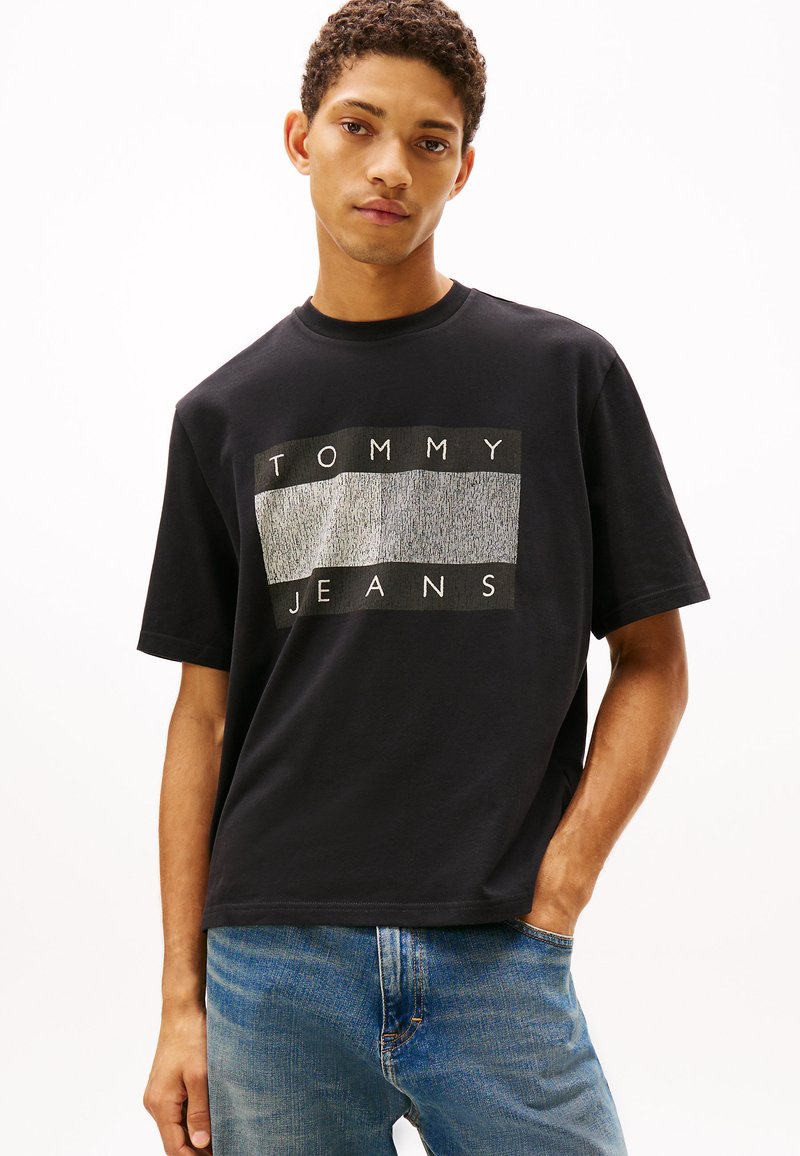 Camiseta negra de algodón con un corte cuadrado, que presenta un logo gráfico "TOMMY JEANS" en gris y blanco en el frente. Combinada con vaqueros de mezclilla azul claro.