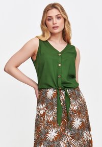 Zielony bezrękawnik z dekoltem w kształcie litery V i drewnianymi guzikami, związany w pasie; zestawiony z brązową wzorzystą spódnicą z białym wzorem liści.