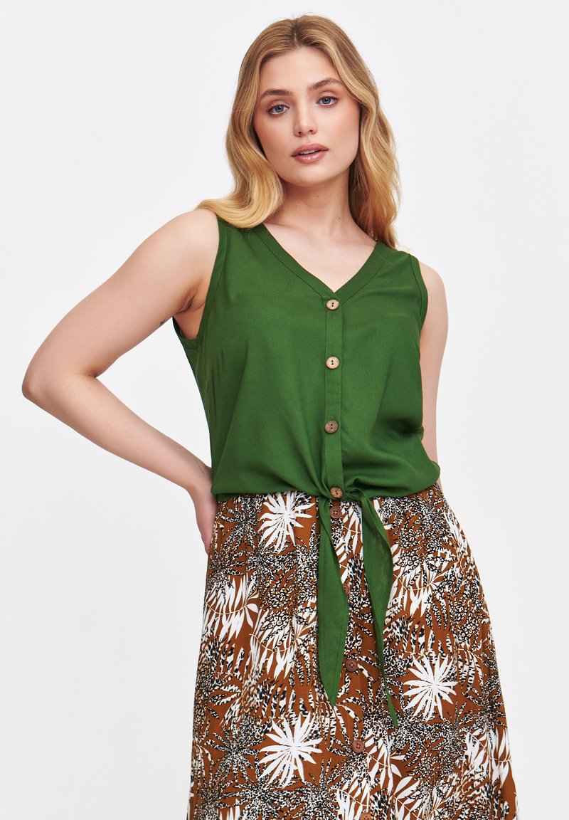 Zielony bezrękawnik z dekoltem w kształcie litery V i drewnianymi guzikami, związany w pasie; zestawiony z brązową wzorzystą spódnicą z białym wzorem liści.