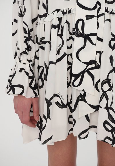 Rochie albă cu un model de fundă neagră, având mâneci lungi cu manșete elastice și o margine în mai multe straturi. Material neted și ușor.