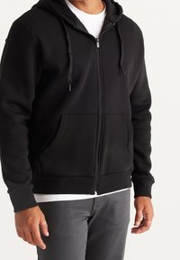 Zwarte zip-up hoodie van zacht materiaal met een twee-paneel capuchon, kangoeroezakken en geribbelde manchetten, waarbij een wit onderhemd zichtbaar is.