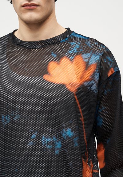 Νεαρό άτομο που φοράει μαύρο διάφανο πουκάμισο με μεγάλο πορτοκαλί λουλούδι και μπλε αφηρημένα σχέδια ορατά από κάτω.