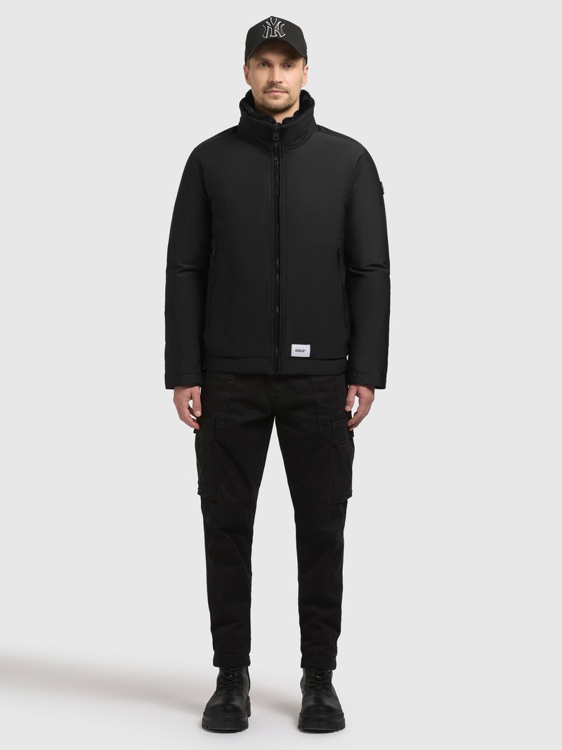 Zwarte gewatteerde jas met hoge kraag en ritssluiting aan de voorkant, gedragen over zwarte cargobroeken en zwarte laarzen. De pet heeft een logo aan de voorkant.