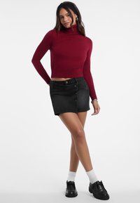 Žena u burgundskom turtleneck puloveru, crnoj mini suknji na dugmad, bijelim čarapama i crnim loafericama, pozira s jednom rukom na boku.