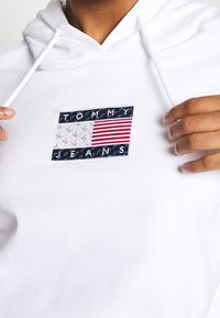 Vit hoodie med en broderad rektangulär Tommy Jeans-logotyp i blått, vitt och rött, som föreställer stjärnor och ränder.