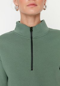 Olivgrünes Zip-up-Oberteil mit hohem Kragen und schwarzem Reißverschluss. Weicher, strukturierter Stoff ohne sichtbare Muster oder Akzente.