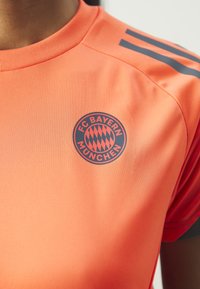 Φανέλα FC Bayern Munich σε πορτοκαλί χρώμα με στρογγυλό λογότυπο που διαθέτει κόκκινο και μαύρο σχέδιο σε διαμάντι, κοντά μανίκια και αντίθετες γκρίζες ρίγες στους ώμους.